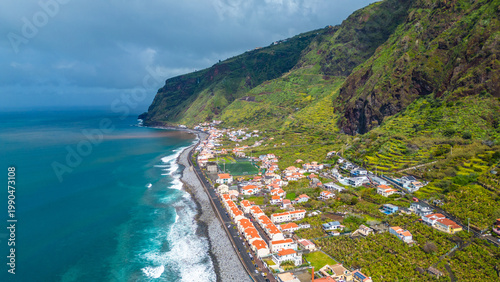 Aerial view of a charming Paul do Mar village nestled beneath lush green cliffs and terraced fields on Madeira Island, Portugal. Atlantic waves meet the pebble beach under an overcast sky.