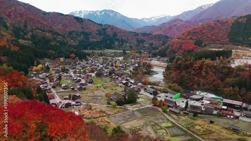 Panoramic drone shot of the historic Shirakawa-go village during autumn. Traditional thatched-roof farmhouses are surrounded by vibrant red and orange foliage with mountains in the background.