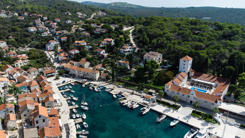 Aerial view of a Mediterranean coastal village with marina, boats and red-roof houses surrounded by green hills and clear turquoise water