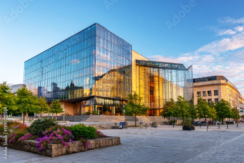 A wide open plaza is seen in front of a big modern building. People walk around the area while enjoying time outside as the sun sets over the city skyline