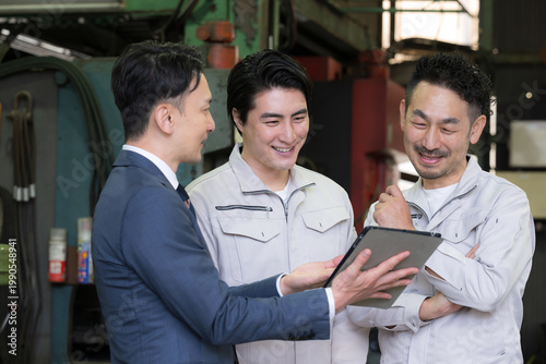 Fotografía 工場でタブレットを使い３人で打ち合わせする作業員とビジネスマンの現場、dxやIT、AI化の機械の導入や信金ビジネスマンが借り入れ提案のイメージ