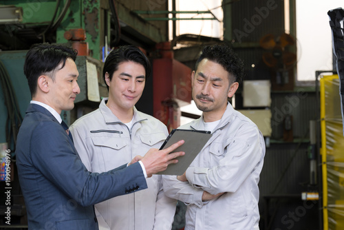 Fotografía 工場でタブレットを使い３人で打ち合わせする作業員とビジネスマンの現場、dxやIT、AI化の機械の導入や信金ビジネスマンが借り入れ提案のイメージ