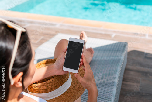 Asian woman using mobile phone while relaxing on outdoor sunbed by the pool at hotel. Work from anywhere, Technology and lifestyle concept. Copy space