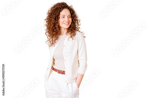 Confident mature woman with red curly hair smiling, looking away, hands in pockets, wearing white casual outfit