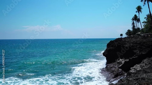 Dramatic Rocky Cliff with Crashing Ocean Waves and Silhouetted Coconut Palms Against Blue Sky - 4K footage
