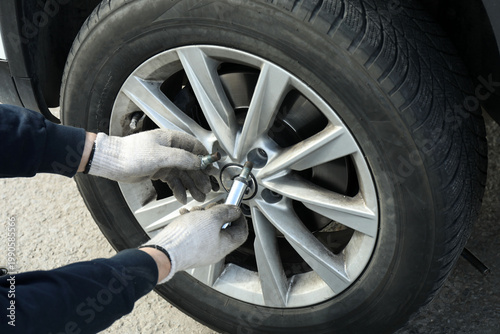 A man in gloves changes a car tire on the road, unscrewing the wheel rim bolts with his fingers, indicating the need for tire replacement or roadside maintenance.
