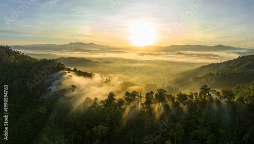 Breathtaking aerial view of the sun rising over a mountain valley covered in a dense sea of rolling clouds. The golden light illuminates the mist, creating a dramatic and ethereal atmosphere at dawn