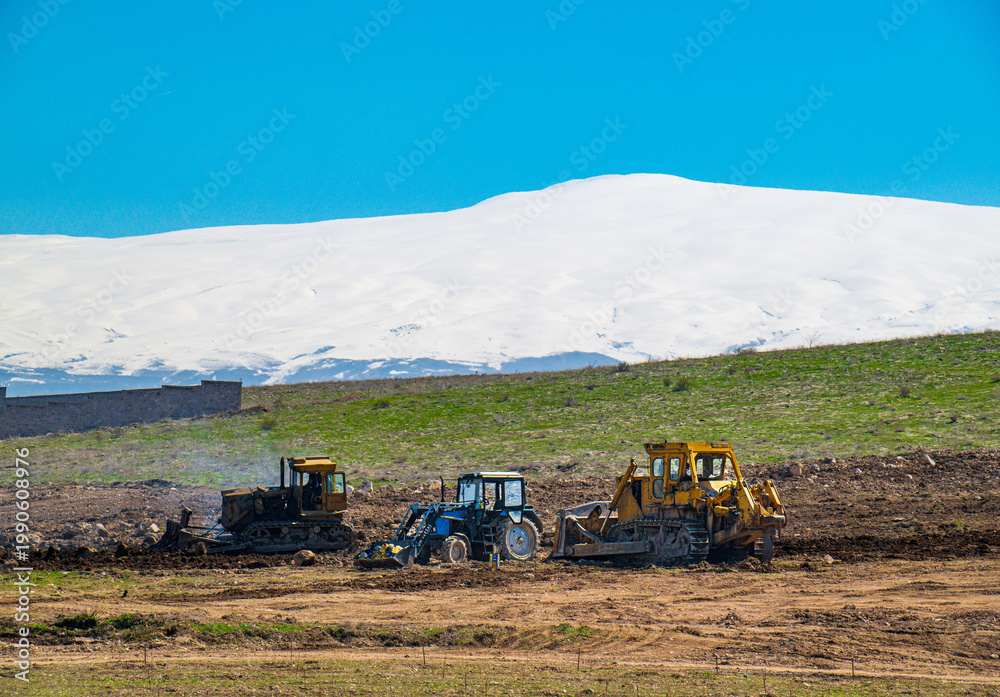 Fototapeta premium Tractors working in field with snowy mountain backdrop