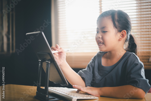 Happy young Asian girl using tablet with keyboard and stylus for online learning, little student smiling during digital classroom session at home office workspace.