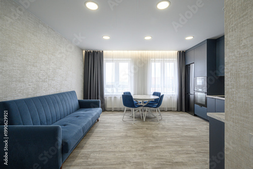 modern living room with a navy blue sofa, round dining table with four blue chairs, dark grey kitchen cabinets, light grey textured walls, sheer white curtains, and recessed ceiling lights
