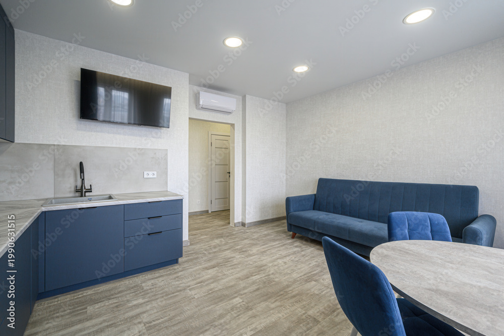 Fototapeta premium modern studio with navy blue sofa and chairs, round dining table, sleek kitchen with dark blue cabinets, wall-mounted TV, air conditioner, light wood flooring, recessed ceiling lights
