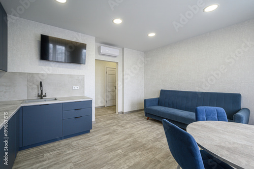 modern studio with navy blue sofa and chairs, round dining table, sleek kitchen with dark blue cabinets, wall-mounted TV, air conditioner, light wood flooring, recessed ceiling lights