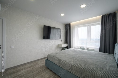 minimalist bedroom with a teal upholstered bed, wall-mounted TV, small white floating desk, grey curtains, large window, light grey textured walls, wooden flooring, and recessed ceiling lights