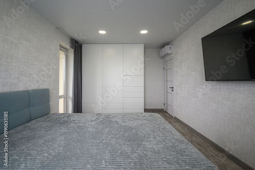 minimalist bedroom with a large white built-in wardrobe, wall-mounted TV, recessed ceiling lights, light grey textured walls, a neatly made bed with a grey quilt, and a small floating shelf.