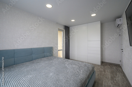 minimalist bedroom with a large white built-in wardrobe, wall-mounted TV, recessed ceiling lights, light grey textured walls, a neatly made bed with a grey quilt, and a small floating shelf.