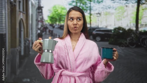 Woman in pink bathrobe holding a stovetop coffee maker and enamel cup, hands visible and puckering lips while standing on a cobblestone street near parked cars and bicycles; morning routine calm.
