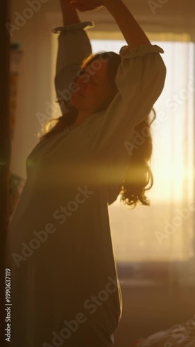 Young pregnant woman waking up at dawn, stretching her arms and tenderly stroking her belly while enjoying the warm morning sunlight streaming through the window of her bedroom