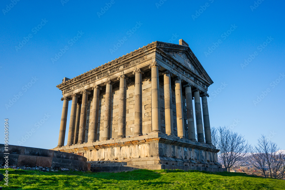 Fototapeta premium Sunlit ancient Garni temple with stone columns, grassy hill, and bare trees