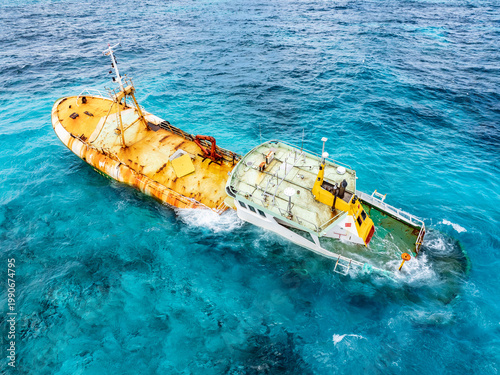 Tablou pe pânză Aerial view of of starboard of a half-sunken fishing boat in inclined position that ran aground on a reef, surrounded by turquoise blue water