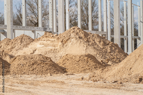 Large piles of sand and aggregate staged on warehouse construction site beside concrete column grid for earthworks, grading, and material supply use