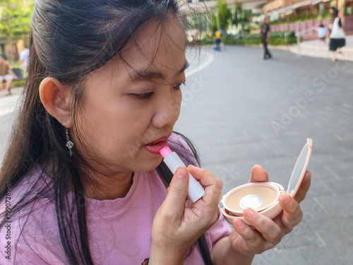 Fényképezés Focused Asian woman outdoors delicately applying vibrant pink tinted lip balm while holding compact powder mirror for a casual beauty touch-up in a public urban setting