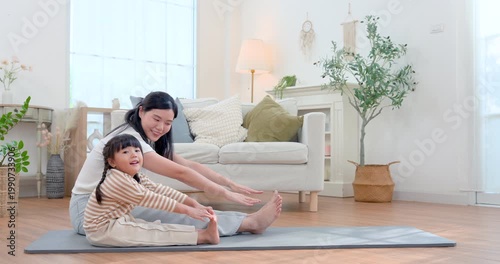 Asian single mother reaches forward on yoga mat beside preschool daughter during playful stretching, encouraging flexibility, body control, active bonding, healthy routine, mindful movement home.