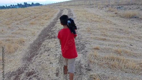 Rear View of Boy Carrying Black Baby Goat on Back Walking in Harvested Field at Sunset