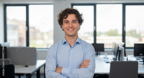 A young man in a light blue shirt stands confidently in a modern office with computers and large windows. His arms are crossed as he smiles at the camera in this bright workspace.
