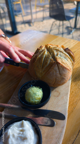 Fresh Crusty Bread and Herb Butter on a Wooden Board Being Sliced with Knife in Restaurant