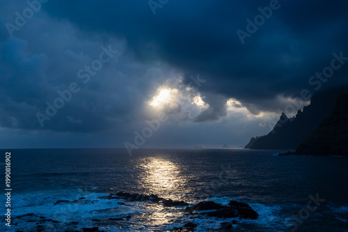 Dark clouds loom over restless ocean waves. Sunlight pierces through thick stormy sky. Rocky shore glows with gentle, scattered light. Stormy sea reflects golden, shimmering beams