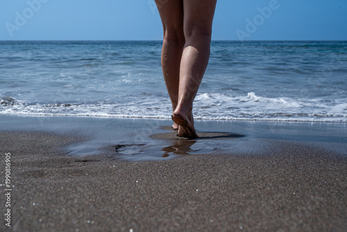 Feet step gently into shallow ocean waves. Sand glistens under soft, sunlit sky. Waves lap at shoreline with quiet rhythm. Person walks barefoot, bare legs visible
