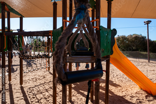 Sunlit playground features a swing and slide. Sand dunes frame the scene beneath a canopy. Children's laughter echoes in the bright air. Ideal for summer, park, or outdoor fun imagery