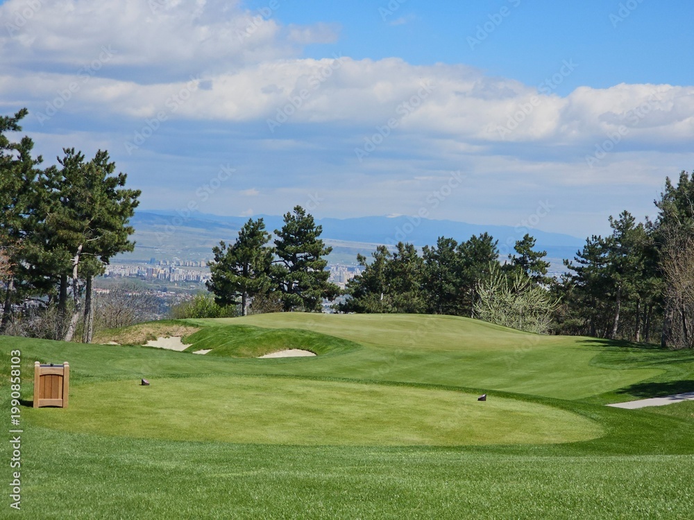Obraz premium Golf course with green fields and trees under a blue sky near a city with mountains