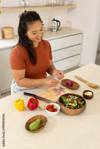 Mid-adult Asian woman preparing salad at kitchen counter in rust top and jeans using wooden board