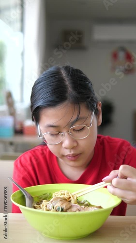 Close-up of young Asian girl with glasses using chopsticks to eat yellow noodle soup in a restaurant, Thai street food and lifestyle concept