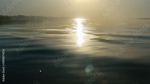 Serene water surface reflecting sunlight at dawn, gentle ripples creating a tranquil scene with distant trees and hazy sky in the background