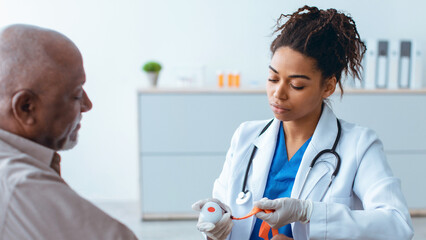 A healthcare worker checks the vital signs of an older man in a medical office. The worker wears a white coat and has a stethoscope. The setting is bright and organized.