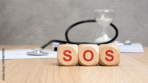 Red SOS word on wooden blocks. Blurred stethoscope and hourglass. Medical emergency concept.