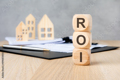 ROI acronym on vertically stacked wooden blocks. Blurred houses and documents. Real estate investment concept.