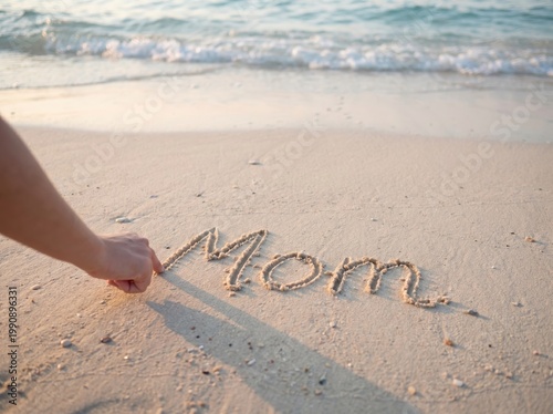 A persons hand writing the word mom in the sand on a beach with waves in the background at sunset