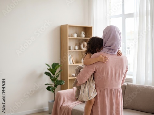 A woman wearing a hijab and a little girl are hugging each other while standing in a bright living room with a couch and a bookshelf