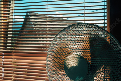 Electric fan in a room by a window with aluminum blinds on a hot summer day.