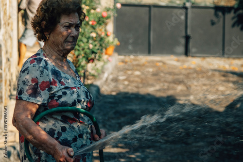 Fotografie Senior woman in floral blouse tends her garden, watering lush green plants with a hose in warm golden-hour light