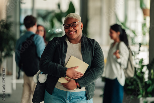 Smiling student holding book and headphones, walking on university campus, embracing education and student life