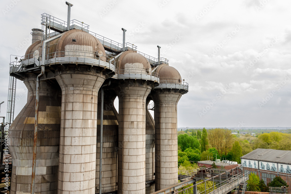 Fototapeta premium Tall industrial towers with dome tops and metal walkway, heavy industry structures overlooking green urban landscape under cloudy sky