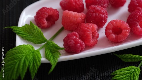 Fresh ripe raspberries with green leaves served on a white ceramic plate over a dark textured background. Close up.