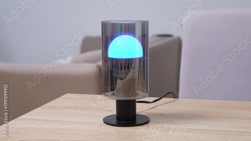 Smart LED lamp with bright blue glowing bulb inside transparent glass cylinder on wooden table indoors, modern smart home technology concept, contemporary interior lifestyle scene.
