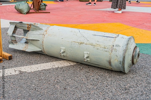 Aged, weathered bomb lying on its side, missing a fin, resting on asphalt or concrete with white parking lines Background has blurred figures and colorful ground design resembling a map or abstract