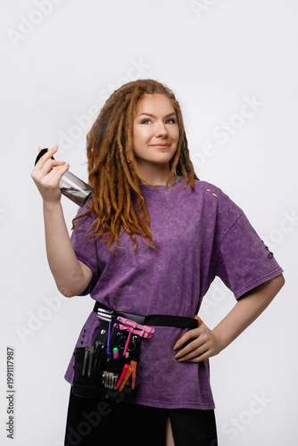 Young Caucasian woman with dreadlocks, wearing purple t-shirt and tools belt, holding spray bottle, smiling, front view, looking at camera. Isolated. Concept hairstyling service, small business.