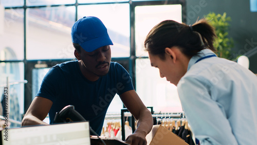 Tablou pe pânză Asian worker signing packages delivery report, discussing clothes distribution with african american courier in modern boutique